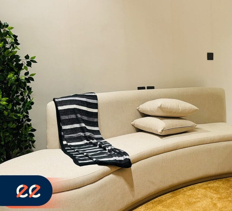 تصوير أريكة بيضاء أنيقة مع وسادتين وغطاء مخطط أسود وأبيض على أحد الأذرع، بجانب نبات أخضر داخلي، مع ديكور حديث لمشاريع إيلفينز.